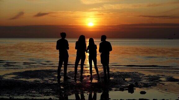 背影剪影的四个年轻的朋友站在海边的海滩上欣赏美丽的橙色日落的水交谈和与啤酒瓶干杯