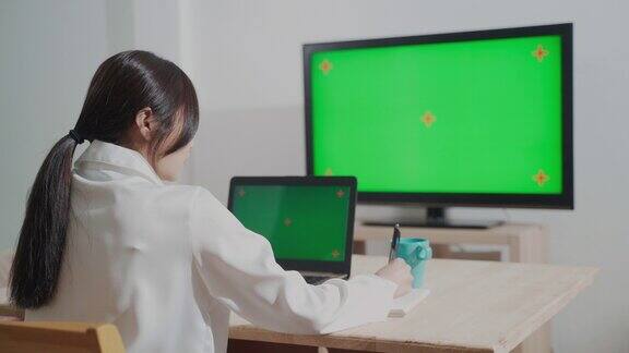 女性使用笔记本电脑和绿屏电视在家工作的后视图