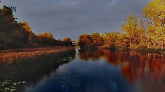 鸟瞰图一架直升机飞过农村河流的黑色水面远处有一座小桥带着金色叶子的树木和芦苇耸立在岸边树木和灰色的天空倒映在平滑的河面上金色的秋天4kProRes