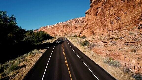 无人机跟随银色汽车在高速公路上行驶在陡峭的峡谷岩石山茂密的沙漠绿化和树木之间