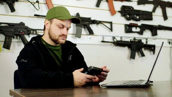 一个枪支贩子在工作坐在商店的电脑前在架子的背景上有不同的武器