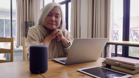 亚洲高级女性在家中客厅使用笔记本电脑和智能音箱工作