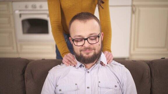 一个戴眼镜的大胡子男人坐在沙发上女人用手按摩他的肩膀一个漂亮的黑发女人弯下腰把头靠在他的肩膀上吻了他的脸颊