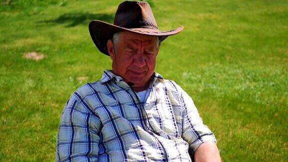 老人累了戴着帽子坐在倾斜的草坪上的椅子上