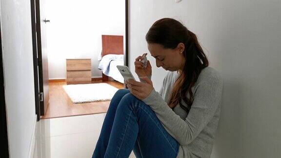 在冠状病毒封锁期间一名沮丧的年轻女子靠墙坐在地板上哭泣看着智能手机