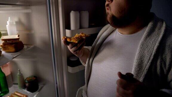 一个懒惰的胖子站在冰箱旁边嚼着披萨喝着啤酒