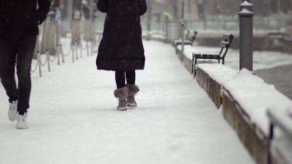 女人的腿毛茸茸的船和黑色的长外套走在雪地覆盖的人行道上在公园在暴风雪期间