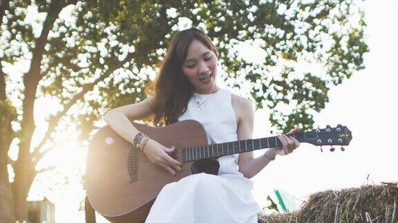 在清晨阳光的照射下一名年轻美丽的亚洲女子穿着白衣在草堆上弹吉他