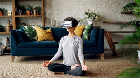 戴着虚拟现实眼镜的苗条女孩正坐在家里的地板上冥想以莲花的姿势享受个人练习现代科技和瑜伽概念