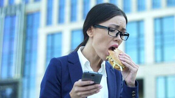 年轻的女经理一边吃着不健康的汉堡一边在手机上查看消息