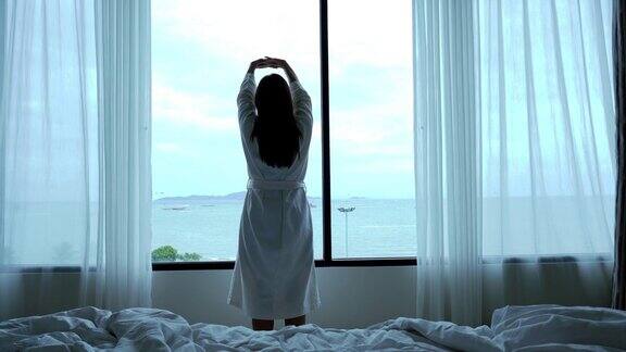 迷人的亚洲女人在早上醒来锻炼看着床上的窗户在豪华酒店卧室生活方式和休闲概念的4k视频的场景背后