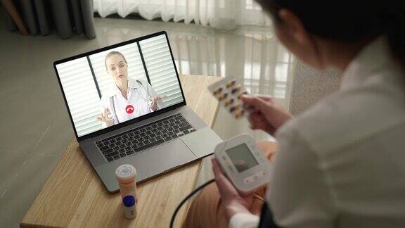 一位亚洲妇女病人在家里用笔记本电脑和她的医生聊天同时使用血压检查