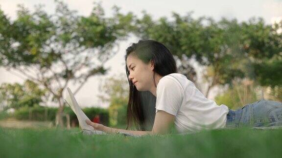 美丽的亚洲女人放松阅读一本书而躺在地板上的草地户外在一个公园在美丽的日落