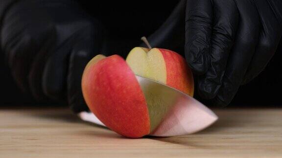 一个红苹果躺在切菜板上一把菜刀把一个红苹果切成两半在黑色的背景上用刀把苹果切成两半人手戴着黑色手套切水果特写慢动作