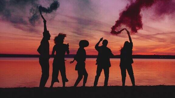 欢乐的朋友们在日落时分手持烟雾弹在湖边跳舞
