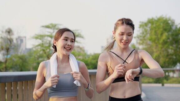 前视图:两个亚洲美丽的中年女性朋友穿着运动服和完美的肌肉一起积极慢跑使用智能手表监测在公园花园的人行天桥靠近中心城市早上积极的生活方式