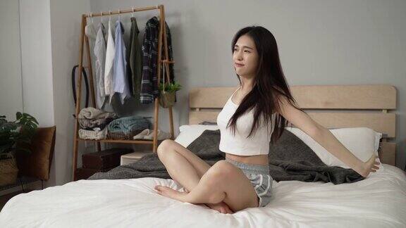 侧面的观点一个漂亮的快乐的亚洲女孩坐在床上看着外面的距离与伸展的感觉在阳光明媚的早晨在家里恢复