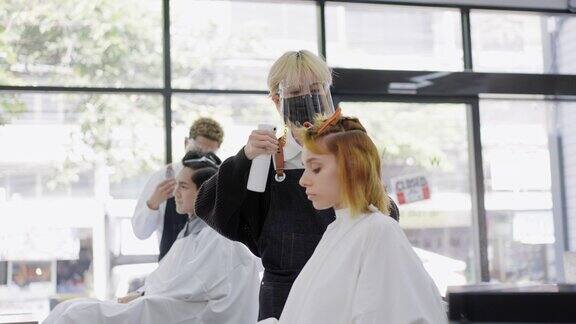专业的亚洲男性造型师戴着防护口罩和面罩在剪头发前对顾客的女性头发进行喷水另一位同事也在打理男性的发型新常态美容院或理发店经营理念