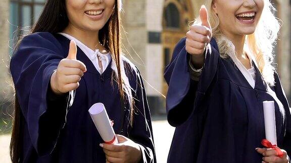 两个美丽的多民族女孩在学术长袍显示拇指向上的标志微笑