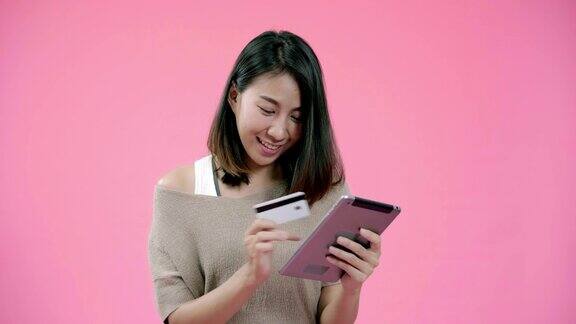 年轻的亚洲女子使用平板电脑购买网上购物信用卡感觉快乐微笑在休闲服装在粉红色的背景工作室拍摄快乐的微笑可爱的快乐的女人庆祝成功