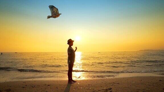 一个年轻男孩的剪影与帽子在一个空的海滩上放风筝背光的夏天日落太阳电影镜头