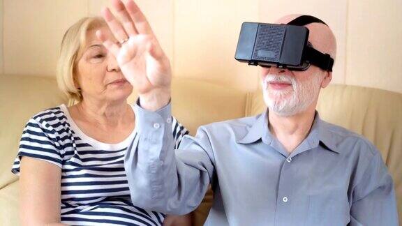 一对愉快、好看的老年夫妇坐在家里的沙发上使用虚拟现实眼镜