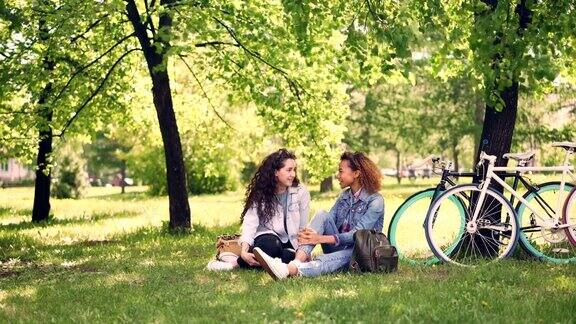 漂亮的白人女孩正在和她的非裔美国朋友聊天在骑完自行车后的休息时间大笑年轻女性坐在公园的草坪上聊天