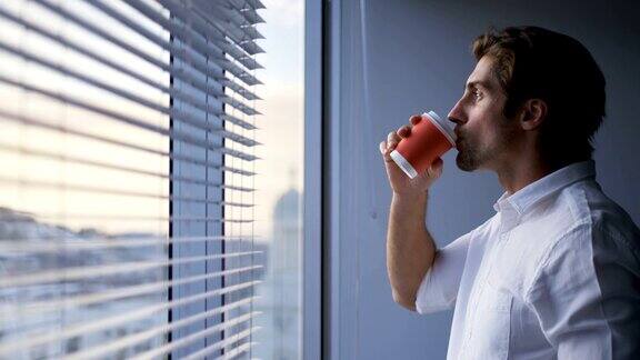 侧面的年轻白人男性主管喝咖啡近窗户在现代办公室4k