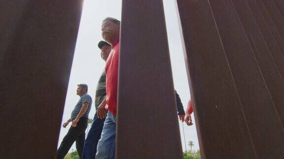 5名西班牙男子和一只狗在美国墨西哥钢条边境墙的墨西哥一侧行走的慢动作拍摄作为摄像机在一个阳光明媚的日子在美国一侧拍摄的电影