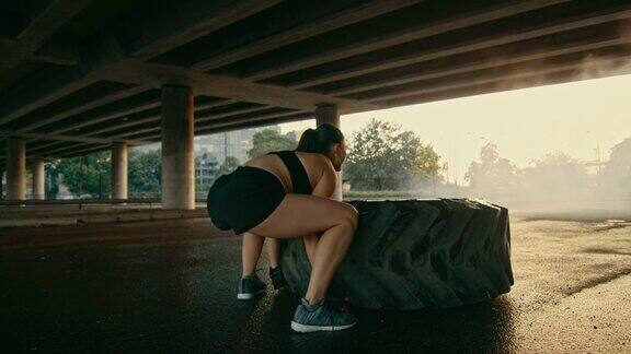 美丽的健身女孩穿着黑色运动上衣和短裤在街上做运动她在城市环境中翻转一个大轮胎