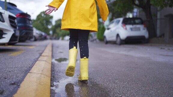 可爱的孩子穿着黄色胶靴和雨衣在街上的水坑里跑