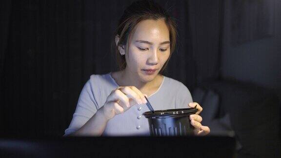 亚洲自由职业者聪明的商业妇女吃方便面而工作在家里的客厅在笔记本电脑在晚上快乐的亚洲女孩坐在桌子上享受放松的时间
