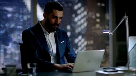 一名商人解开夹克坐在办公桌前开始在笔记本电脑上打字他的办公室可以俯瞰整个大城市