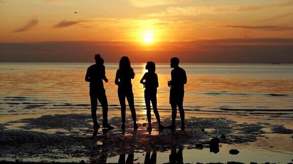 背影剪影的四个年轻的朋友站在海边的海滩上欣赏美丽的橙色日落的水喝啤酒和聊天