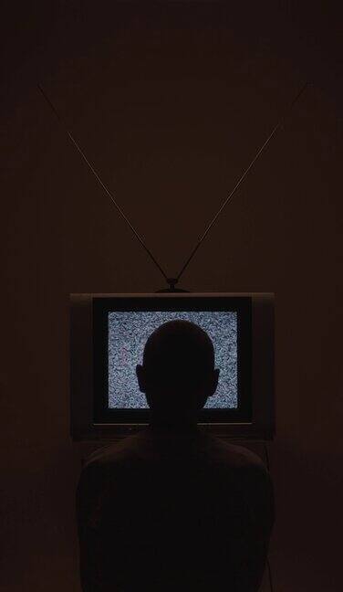 一个垂直的视频一个男人走进房间坐在电视机前电视机的静电噪音在黑暗的灯光下的画面中央