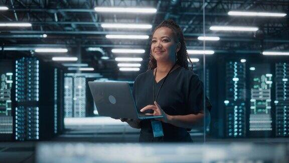 成功的非洲裔美国女性IT专家使用笔记本电脑的肖像站在数据中心系统管理员工作在Web服务云计算服务器分析网络安全