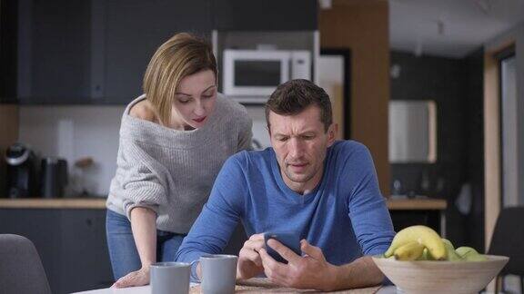 丈夫一边用智能手机上网一边被坐在厨房桌旁的妻子的谈话分心不满的白人男人对呆在家里的女人感到恼怒