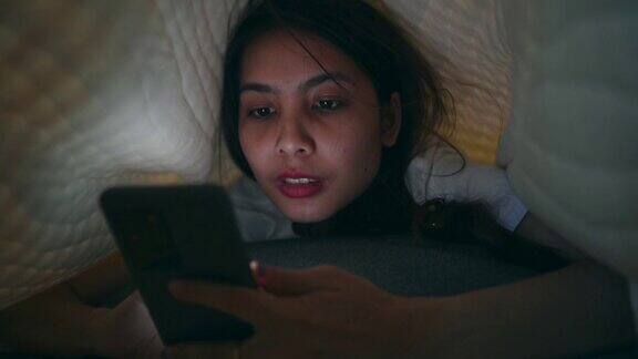 年轻的亚洲女性睡觉时盖着毯子玩智能手机