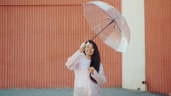 快乐的亚洲女孩穿着雨衣在户外跳舞拿着雨伞看着相机