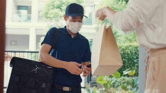 年轻的亚洲快递员戴着面具穿着蓝色海军制服携带箱子敲门回家网上购物纸袋到女人在门前女士扫描二维码支付在家里的智能手机