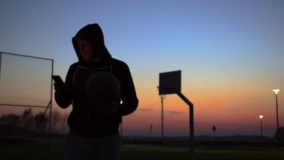 年轻男子使用智能手机在黄昏时走出户外篮球场