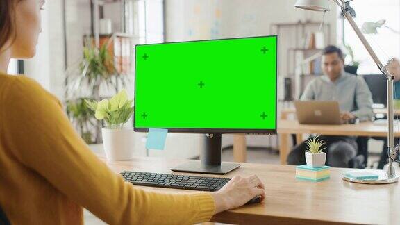 过肩:有创意的年轻女子坐在她的办公桌上使用电脑与模拟的绿色屏幕在背景明亮的办公室多元化的年轻专业团队工作