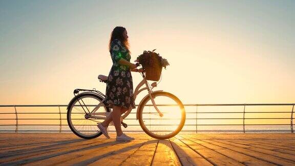 年轻女子的剪影穿着复古自行车和鲜花走在海边的木堤岸在日出或日落浪漫的旅行概念美丽的景色