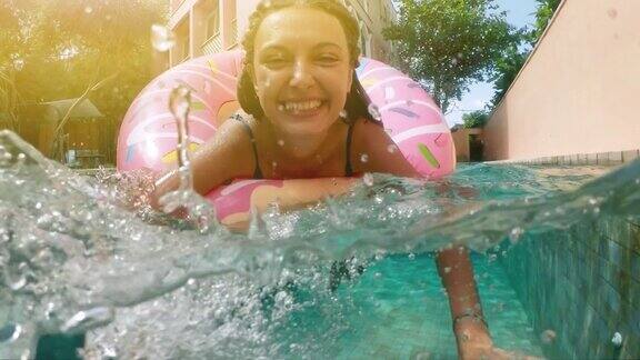 在阳光明媚的夏日里一个戴着太阳镜穿着蓝色比基尼的女人躺在充气的粉色甜甜圈里漂浮在游泳池里看相机女子比基尼游泳池在西瓜橡胶圈放松度假