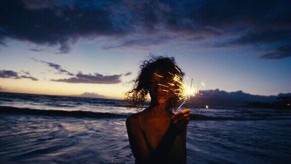 黄昏时分美丽的年轻女子手拿闪亮的烟花走在海滩上