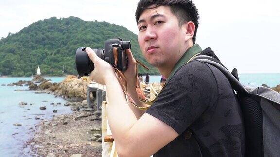 年轻的亚洲男子摄影师微笑和拍照热带岛屿海滩海景背景暑假和度假旅游