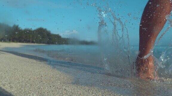 慢镜头:当一名男子沿着海滩奔跑时玻璃般的水珠在他的脚周围飞舞