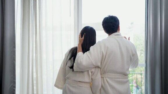 年轻的亚洲夫妇在早上醒来时穿着浴袍打开窗户上的窗帘在豪华酒店的卧室