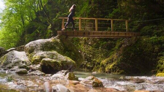 一位年轻的女游客在阳光明媚的日子徒步穿越山溪峡谷上的木桥