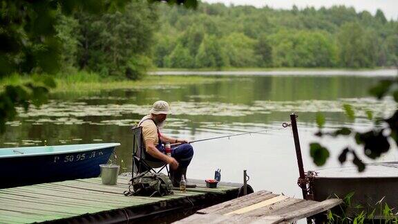 在乡村湖面上的木墩上一名戴着水桶帽的大胡子老人坐在椅子上钓鱼、喝啤酒
